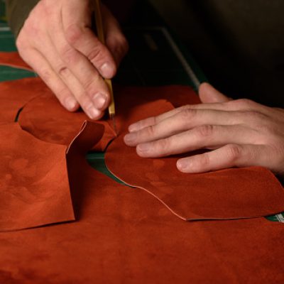 Shoemaker's hands holding craft knife cutting suede leather for handmade shoes on his workplace in workshop. Closeup
